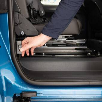 A hand pressing the control switch for the BraunAbility Caroslide Powered Seat Runner on the side of a car's doorframe to move the seat forward or backwards within the car.
