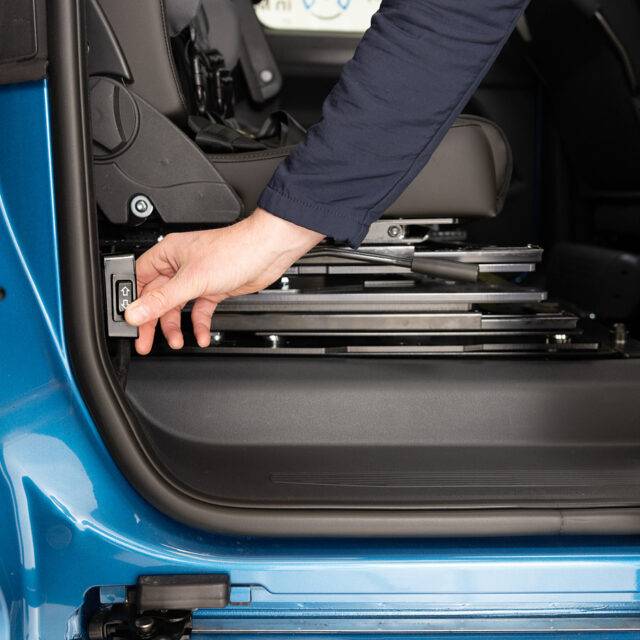 A hand pressing the control switch for the BraunAbility Caroslide Powered Seat Runner on the side of a car's doorframe to move the seat forward or backwards within the car.