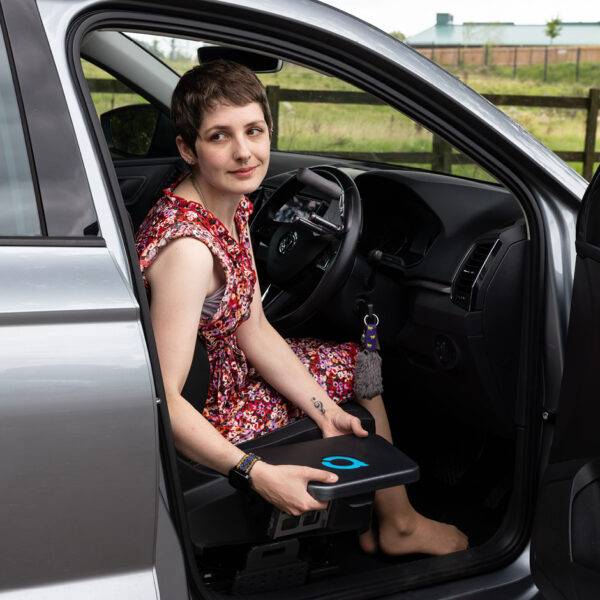 A young woman sat in the driver's seat of her car holding the Autochair Transfer Plate. Positioned next to the seat of the car is a mounting point for the plate.