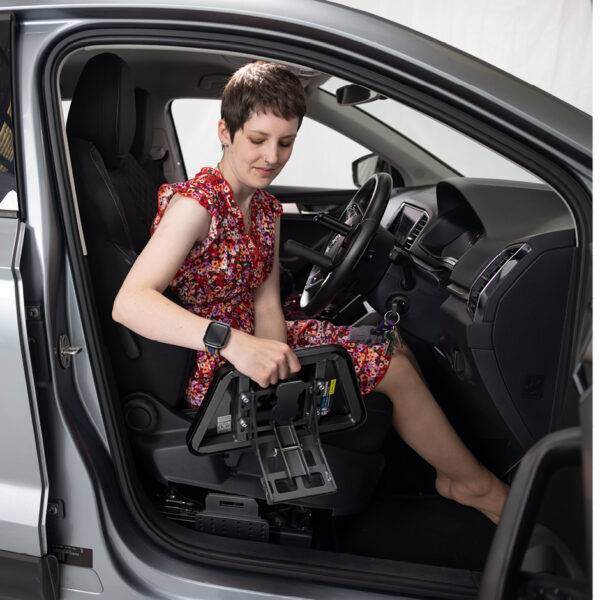 A young woman sat in the driver's seat of her car holding the Autochair Transfer Plate. Positioned next to the seat of the car is a mounting point for the plate.