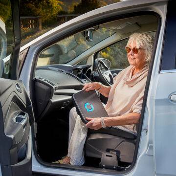 An older woman sat in the passenger seat of her car holding the Autochair Transfer Plate. Positioned next to the seat of the car is a mounting point for the plate.