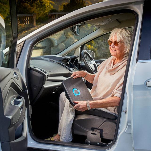 An older woman sat in the passenger seat of her car holding the Autochair Transfer Plate. Positioned next to the seat of the car is a mounting point for the plate.