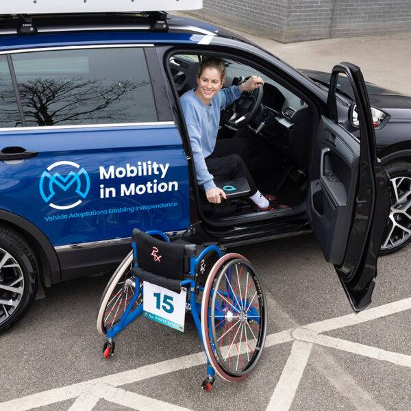 Parlympian Robyn Love sat in the driver's seat of a Mobility in Motion Demo Car. The Autochair Transfer Plate, a small black removable platform, is positioned next to the driver's seat of the car. She is leaning on the platform to get closer to her wheelchair.