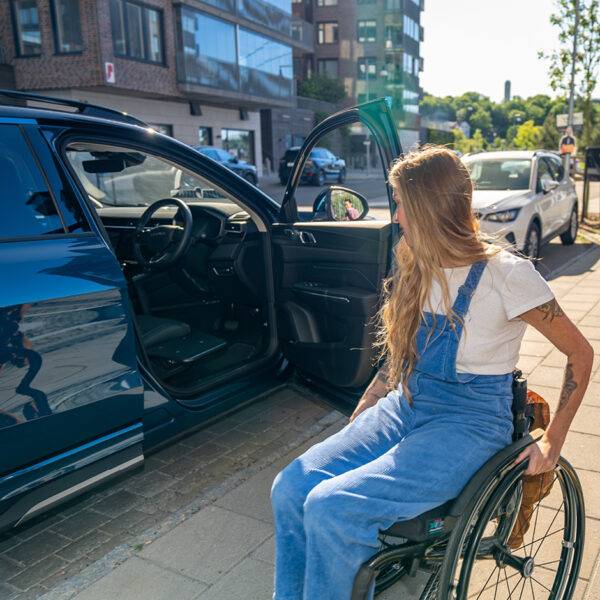 A young woman in a wheelchair looks toward her car's open door. The BraunAbility Transfer Board, a small black folding platform, is positioned next to the driver's seat of the car.