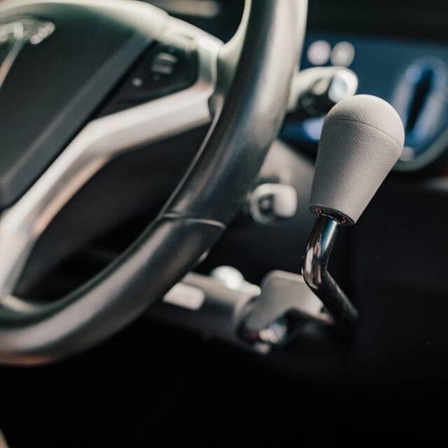 a driver's seat view of the steering wheel and the Veigel eConnect positioned beside it. It is a modern and sleek black push/pull electronic hand control with a sleek rounded handle.