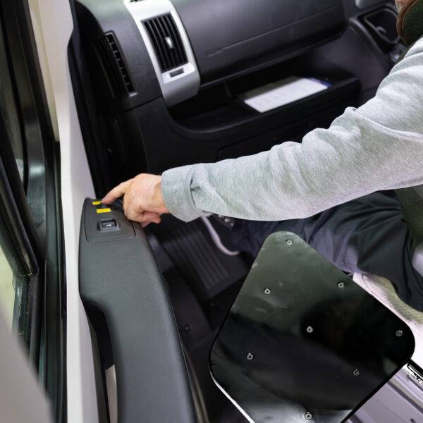 In the passenger seat of a car, a man is pressing the up/down control for an electric transfer plate positioned next to the seat.