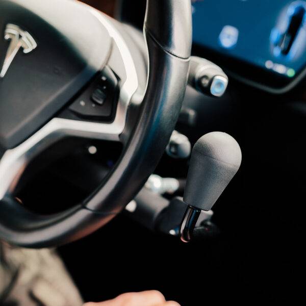 a driver's seat view of the steering wheel and the Veigel eConnect positioned beside it. It is a modern and sleek black push/pull electronic hand control with a sleek rounded handle.