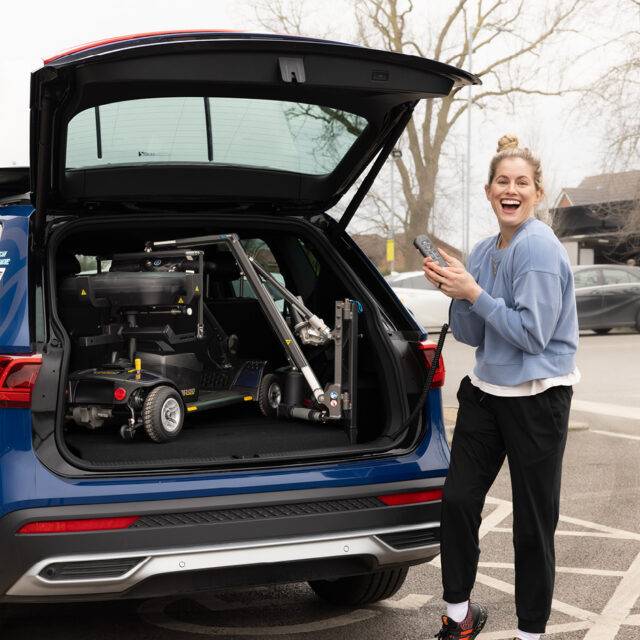 Paralympian Robyn Love at the open boot of a car with a boot hoist lifting a mobility scooter. The scooter has the Autochair Smart Scooter seat installed. It has had the arms and backrest removed so it can be lifted more easily and fit the vehicle.