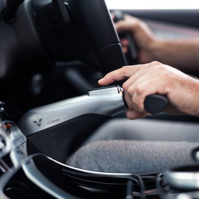 A view of a driver holding the Veigel eClassic hand control, a silver and black twist & push handle hand control for accelerating and braking their car. They are pressing the button on top of the handle to hold the brake.