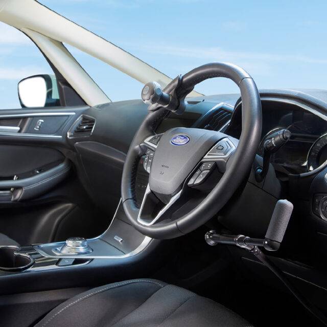 A driver's seat view of the wheel in a car with mechanical push/pull hand controls next to the steering wheel and a steering ball on the wheel.