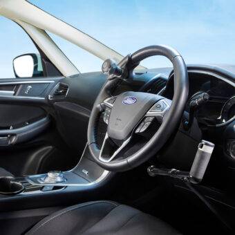 A driver's seat view of the wheel in a car with mechanical push/pull hand controls next to the steering wheel and a steering ball on the wheel.