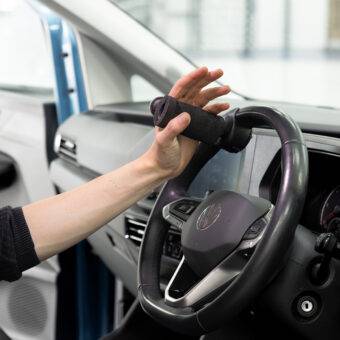 A driver's seat view of the steering wheel. The driver is holding the wheel via the BraunAbility 2-Pin Specialist Steering Aid. It is a soft foam handle perpendicular to the wheel with a supportive pin to hold the hand in place.