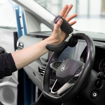 A driver's seat view of the steering wheel. The driver is holding the wheel via the BraunAbility 3-Pin Specialist Steering Aid. It is a soft foam handle perpendicular to the steering wheel with a supportive channel and two wrist pins to hold the hand and wrist comfortably in place. It has a separate mounting adapter that connects to the steering wheel allowing the 3-Pin grip to be easily removed.