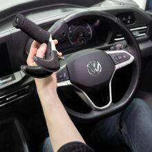 A driver's seat view of the steering wheel. The driver is holding the quick-release specialist steering aid the BraunAbility 3-Pin Specialist Steering Aid. It is a soft foam handle perpendicular to the steering wheel with a supportive channel and two wrist pins to hold the hand and wrist comfortably in place. It has a separate mounting adapter that connects to the steering wheel allowing the 3-Pin grip to be easily removed.
