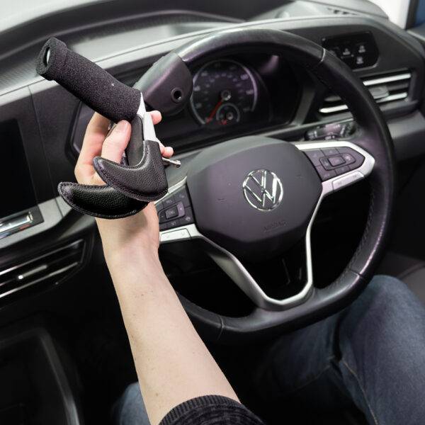 A driver's seat view of the steering wheel. The driver is holding the quick-release specialist steering aid the BraunAbility 3-Pin Specialist Steering Aid. It is a soft foam handle perpendicular to the steering wheel with a supportive channel and two wrist pins to hold the hand and wrist comfortably in place. It has a separate mounting adapter that connects to the steering wheel allowing the 3-Pin grip to be easily removed.