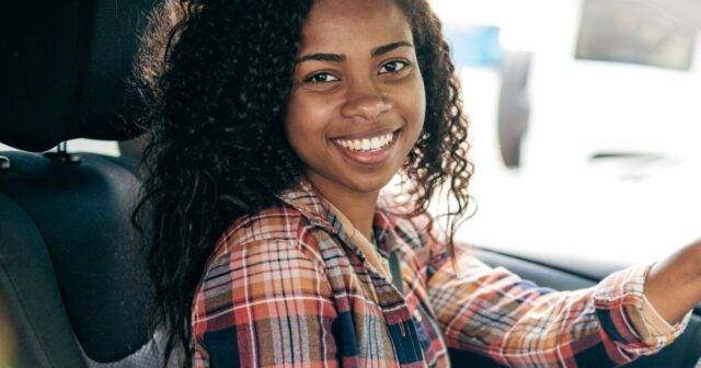 Young woman sat in driver seat smiling wearing a tartan shirt.