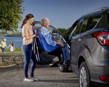 Adult daughter helping to transfer her elderly dad into the car using a person hoist