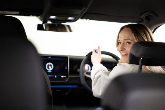 Young woman sat in drivers seat looking over her left shoulder smiling. Hand on secondary remote control steering device.