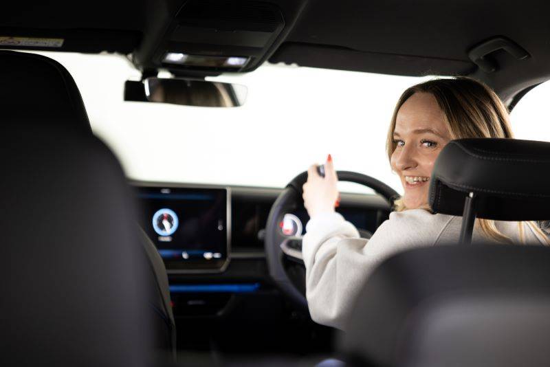 Young woman sat in drivers seat looking over her left shoulder smiling. Hand on secondary remote control steering device.