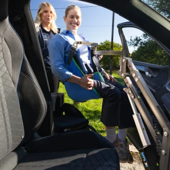 A Mobility in Motion team member operates the MPL XR person hoist to transfer Robyn Love in a wheelchair into a car, viewed from inside the vehicle.