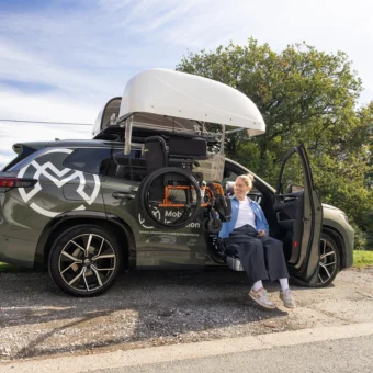Robyn Love sat on a swivel seat fitted in a parked vehicle in a sunny country park, facilitating a safe and comfortable transfer. She also operates the wheelchair roof box to lift and load her folding wheelchair.